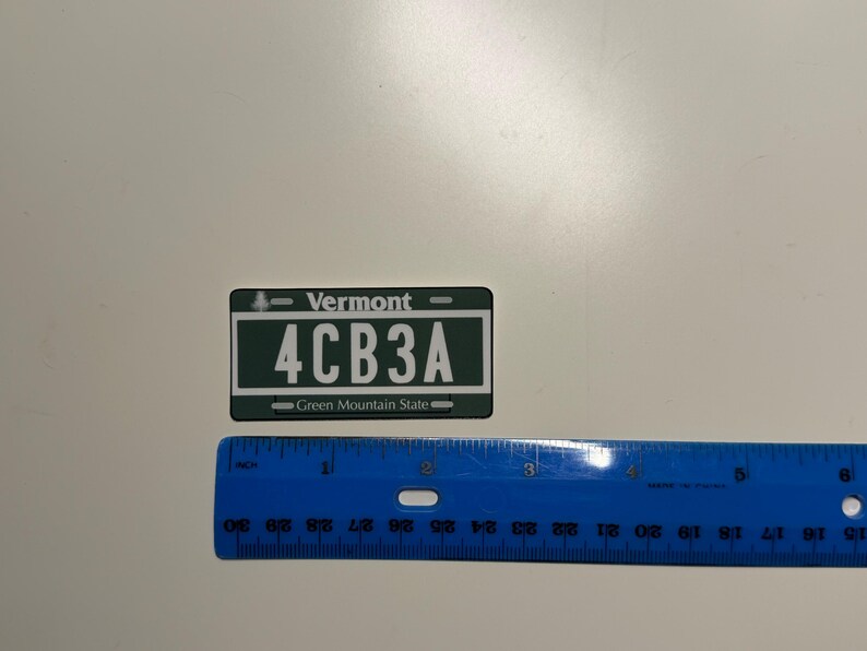 May include: A rectangular Vermont license plate replica in dark green and white, featuring the text Vermont, 4CB3A, and Green Mountain State. A blue plastic ruler is positioned below the plate, showing it measures approximately 3.5 inches in width.