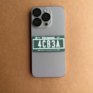 May include: A light gray smartphone with a triple-lens camera system featuring a rectangular sticker on the back. The sticker is a green Vermont license plate replica with white text reading Vermont, 4CB3A, and Green Mountain State.