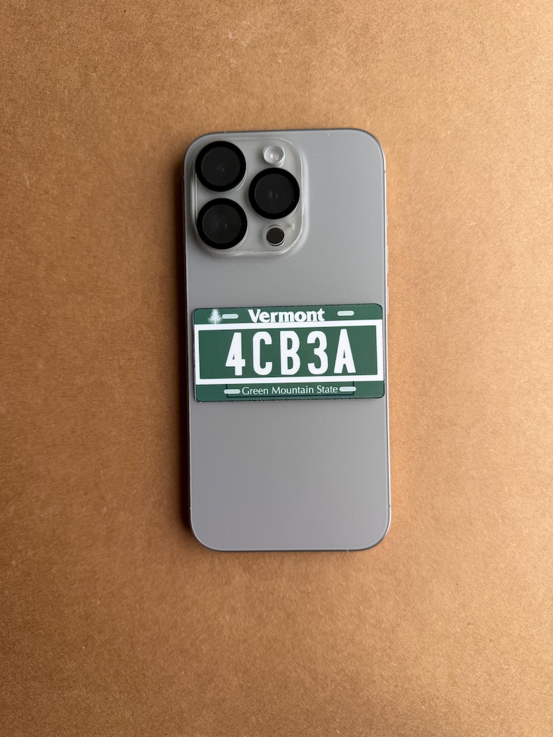 May include: A light gray smartphone with a triple-lens camera system featuring a rectangular sticker on the back. The sticker is a green Vermont license plate replica with white text reading Vermont, 4CB3A, and Green Mountain State.