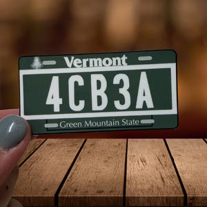 May include: A rectangular Vermont license plate replica with a dark green background and white text. The plate features the word Vermont at the top, the alphanumeric code 4CB3A in the center, and the phrase Green Mountain State at the bottom.
