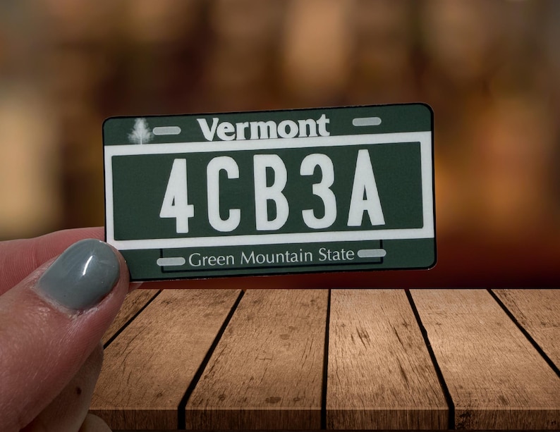 May include: A rectangular Vermont license plate replica with a dark green background and white text. The plate features the word Vermont at the top, the alphanumeric code 4CB3A in the center, and the phrase Green Mountain State at the bottom.