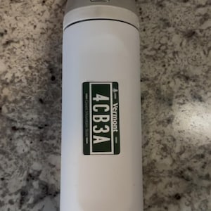 May include: A white cylindrical water bottle with a gray flip-top lid. A rectangular green sticker is centered on the body, featuring the text 4CB3A, the word Vermont, and the phrase Green Mountain State in white lettering on a dark green background.