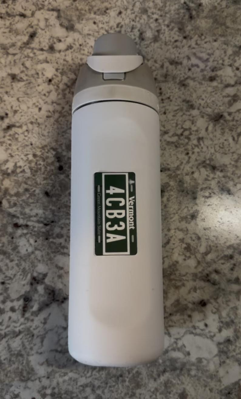 May include: A white cylindrical water bottle with a gray flip-top lid. A rectangular green sticker is centered on the body, featuring the text 4CB3A, the word Vermont, and the phrase Green Mountain State in white lettering on a dark green background.