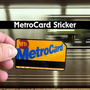 May include: A MetroCard sticker with the text "MetroCard" in blue, on a yellow background, with the MTA logo. The sticker is held in front of a blurred background of a train. The text "Insert this way / This side facing you" is also visible.