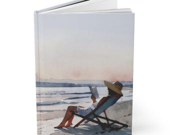 May include: Hardcover journal featuring a watercolor illustration of a person relaxing on a beach. The image depicts a person in a beach chair reading a book, with a hat on, overlooking the ocean. The journal has a white spine and pages.