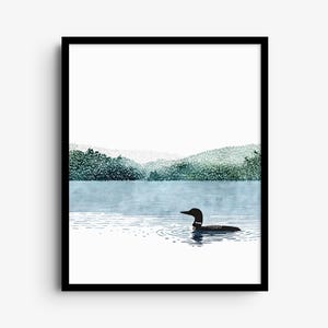 May include: A framed wall art print featuring a black loon swimming on a calm blue lake. The background displays a distant shoreline with green forest hills under a white sky. The artwork is enclosed in a simple black rectangular frame.