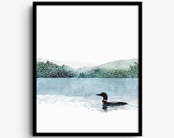 May include: A framed wall art print featuring a black loon swimming on a calm blue lake. The background displays a distant shoreline with green forest hills under a white sky. The artwork is enclosed in a simple black rectangular frame.