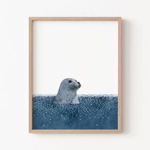 May include: A framed art print showing a watercolour illustration of a seal in blue water. The seal is grey with dark spots, and the water is a deep blue with white speckles. The frame is a light wood colour.