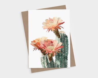 May include: A greeting card featuring a watercolor illustration of a green cactus with three large, blooming flowers in shades of peach, coral, and pink. The cactus has visible spines and is set against a plain white background.