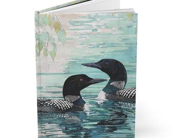 May include: A hardcover journal featuring a watercolor painting of two loons on a lake. The birds have black heads and white-speckled bodies. The background shows a serene lake scene with green and blue hues.