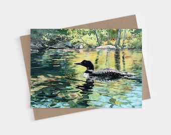 May include: A greeting card featuring a watercolor painting of a black and white loon swimming on a calm lake. The water reflects green and yellow forest foliage, creating ripples around the bird. The card is placed on a brown envelope.