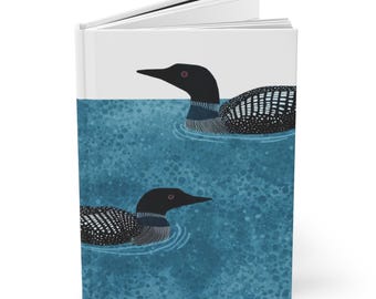 May include: A hardcover journal featuring an illustration of two loons swimming on a blue water background. The loons have black heads, white-speckled bodies, and red eyes. The journal's cover is white with a clean, modern design.