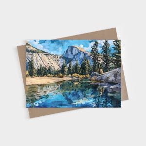 May include: A rectangular greeting card featuring a painterly illustration of a mountain landscape with a granite dome peak, a dense forest of pine trees, and a calm body of water in the foreground reflecting the blue sky and rocky terrain.