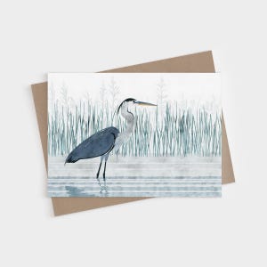 May include: A greeting card featuring a watercolor illustration of a great blue heron standing in shallow water. The bird has grey and blue plumage with a long neck and yellow beak. Tall green reeds are visible in the background.
