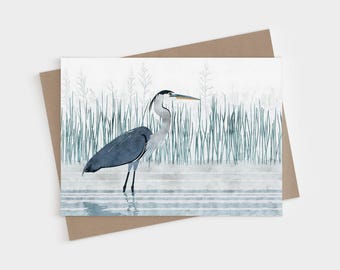 May include: A greeting card featuring a watercolor illustration of a great blue heron standing in shallow water. The bird has grey and blue plumage with a long neck and yellow beak. Tall green reeds are visible in the background.