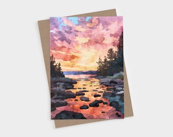May include: A rectangular greeting card featuring a watercolor painting of a sunset over a rocky shoreline. The sky is filled with pink, orange, and yellow clouds above dark silhouettes of pine trees and water reflecting the warm light.