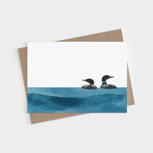 May include: A rectangular greeting card featuring an illustration of two loons swimming in deep blue water with white speckles. The loons have black heads, red eyes, and white-spotted bodies. The card is placed on a brown envelope.