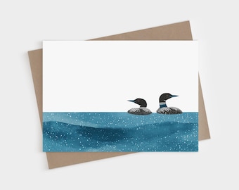 May include: A rectangular greeting card featuring an illustration of two loons swimming in deep blue water with white speckles. The loons have black heads, red eyes, and white-spotted bodies. The card is placed on a brown envelope.
