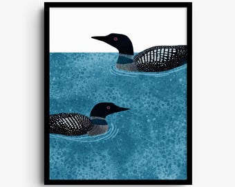 May include: A framed art print featuring two black and white loons swimming in textured blue water. The birds have spotted plumage, dark heads, and red eyes. The background consists of a solid white upper section and a mottled blue lower section.