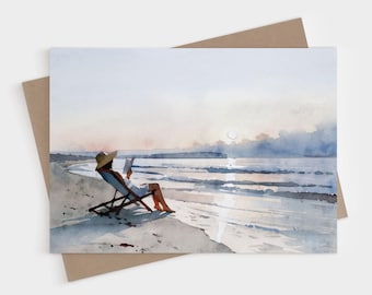 May include: A watercolor painting depicts a figure wearing a wide-brimmed straw hat, sitting in a wooden beach chair on the sand, reading a book. The scene captures a calm ocean shoreline at sunset with soft blue, gray, and pale orange hues.