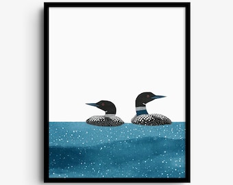 May include: A framed art print featuring two loons swimming in blue water. The loons have black heads, red eyes, and white-spotted black bodies. The water is depicted in shades of blue with white speckles, set against a plain white background.