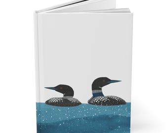 May include: A hardcover journal featuring a serene illustration of two loons floating on blue water. The loons have black heads, red eyes, and white-speckled bodies. The water has a textured appearance with white dots.