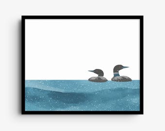 May include: A framed wall art print featuring two loons swimming in blue water. The loons have black heads, red eyes, and white-spotted bodies. The water is depicted with a textured blue watercolor effect and small white dots.