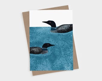 May include: A greeting card featuring an illustration of two black and white loons swimming in textured blue water. The loons have red eyes and detailed plumage patterns. The card is placed on top of a plain brown envelope.