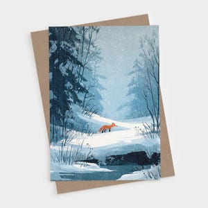 May include: A rectangular greeting card featuring a winter forest illustration with a small orange fox standing in deep white snow near a dark blue stream, surrounded by tall pine trees under a light blue sky with falling snow, paired with a brown envelope.