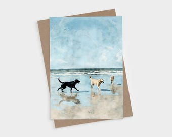 May include: A rectangular greeting card featuring a watercolor illustration of three dogs playing on a sandy beach. A black dog and two yellow dogs are depicted near the ocean waves under a light blue sky with soft clouds.