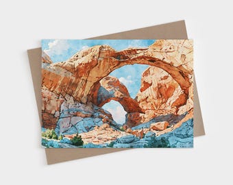 May include: A watercolor painting of a double natural sandstone arch formation in a desert landscape. The rock structures are shades of orange, red, and tan, set against a bright blue sky with white clouds and blue-toned shadows below.