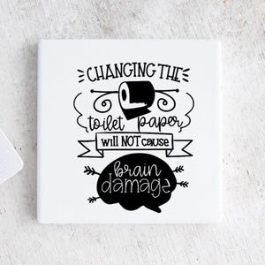 May include: White square coaster with black text that reads "Changing the toilet paper will not cause brain damage". The text is in a handwritten style with a cartoon toilet paper roll and a brain graphic.