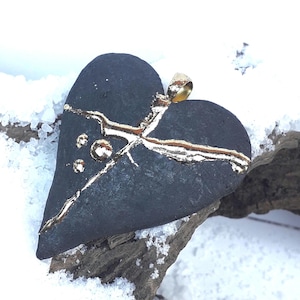 Op de afbeelding: Een donkergrijze hartvormige hanger met goudkleurige accenten. De hanger heeft een gebarsten ontwerp met goud dat de scheuren opvult en drie kleine gouden cirkels. De hanger ligt op een stuk hout met sneeuw op de achtergrond.