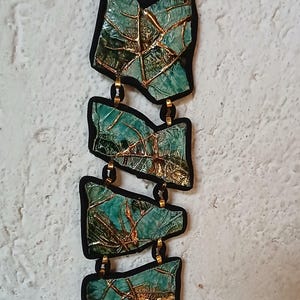 Op de afbeelding: Een verticale ketting met vier onregelmatige, blauwgroene segmenten met een getextureerd, gebarsten oppervlak en metallic gouden aders. Elk segment is zwart omlijnd en verbonden door kleine goudkleurige metalen ringen aan een zwart koord.