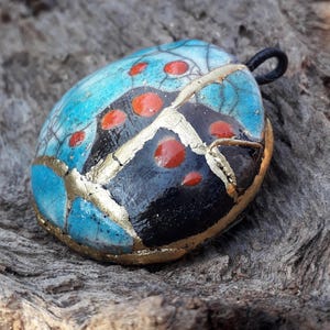 Op de afbeelding: Een blauwe, zwarte en gouden keramische hanger met rode stippen. De hanger is gevormd als een lieveheersbeestje en heeft een zwarte lus om op te hangen.