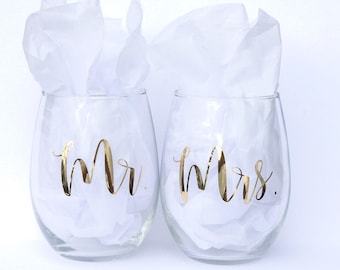 May include: Two clear glass wine glasses with gold lettering. The glass on the left says "Mr." and the glass on the right says "Mrs."