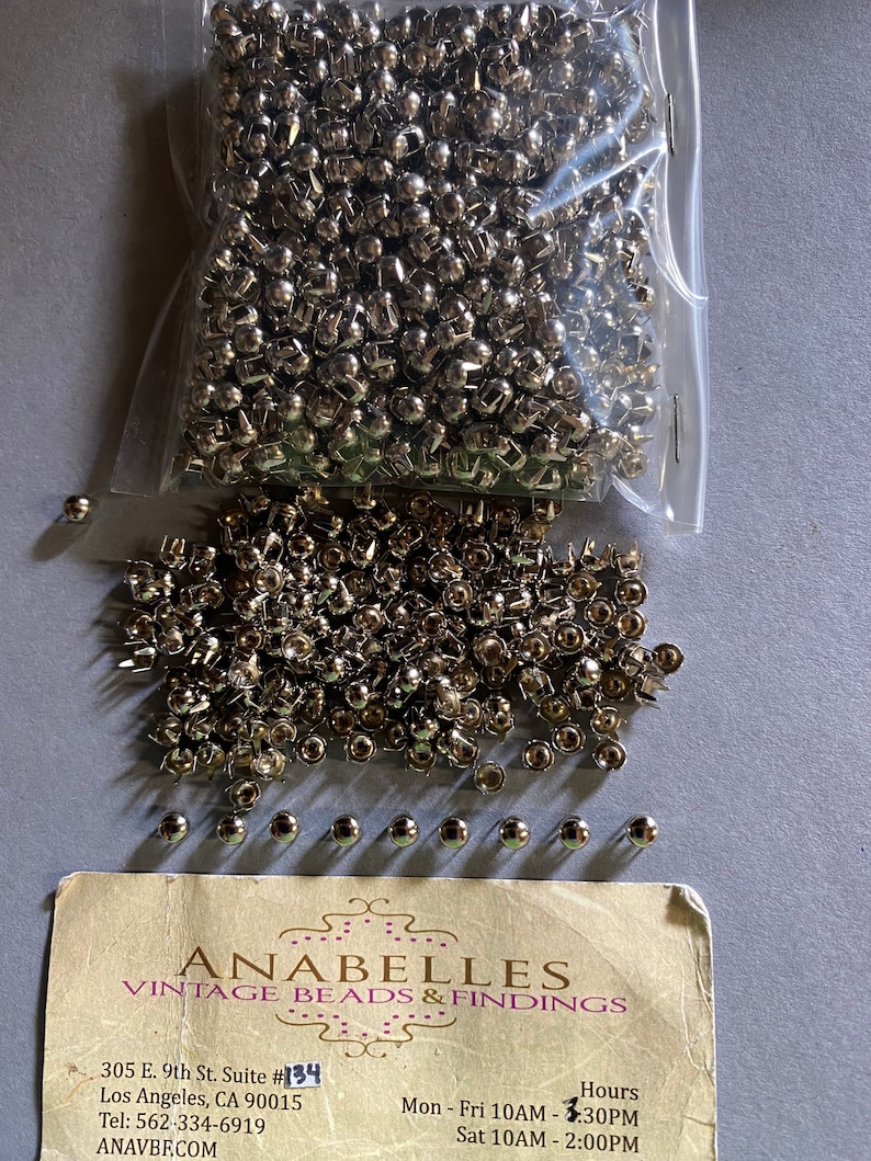 May include: A bag of silver metal rivets, with a few scattered on a white surface. The bag is labelled "Anabelles Vintage Beads & Findings" with the address "305 E. 9th St. Suite #134 Los Angeles, CA 90015 Tel: 562-334-6919 ANAVBE.COM" and the hours "Mon-Fri 10AM-3:30PM Sat 10AM - 2:00PM".