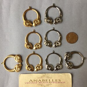 May include: A collection of gold and silver-toned ram head pendants, arranged on a gray surface. The pendants are circular with detailed ram head designs. A business card for Anabelles Vintage Beads & Findings is also present.