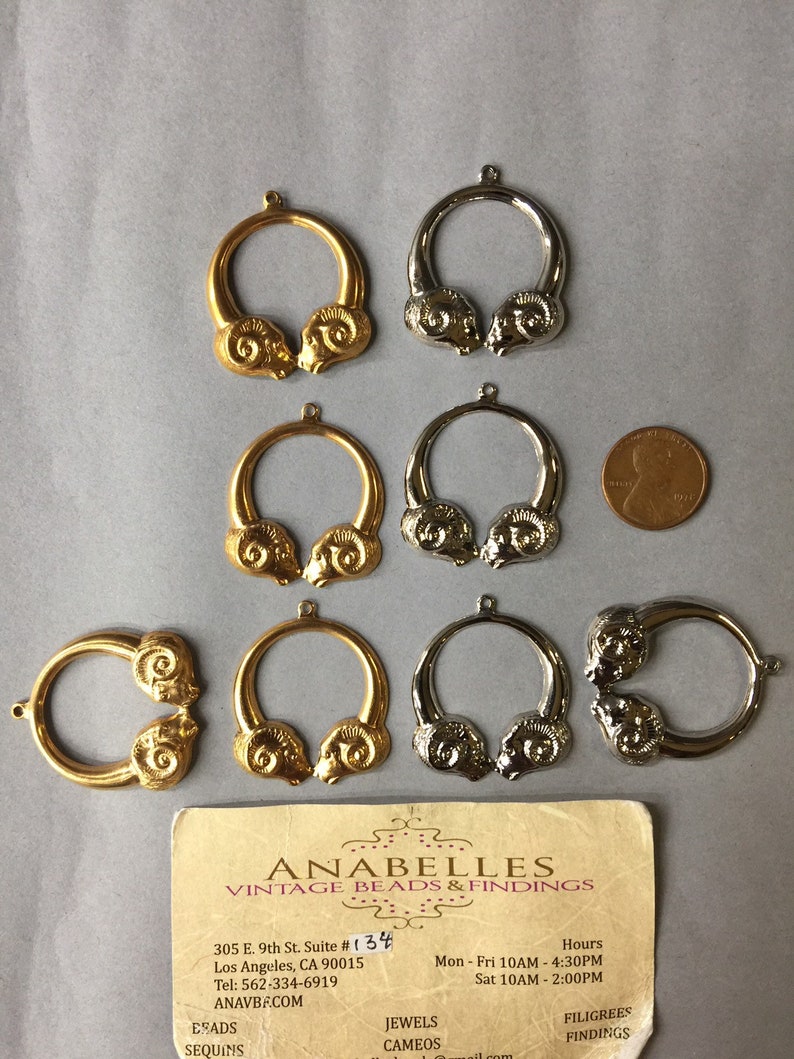 May include: A collection of gold and silver-toned ram head pendants, arranged on a gray surface. The pendants are circular with detailed ram head designs. A business card for Anabelles Vintage Beads & Findings is also present.