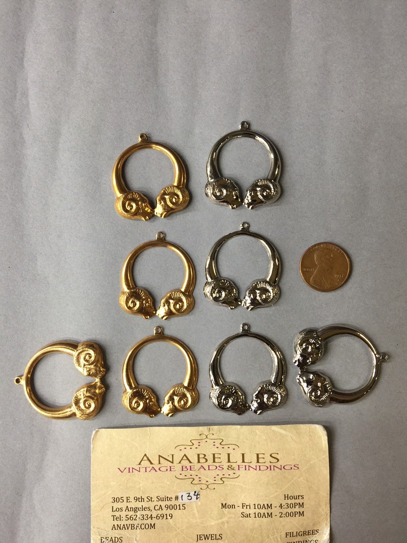 May include: A collection of gold and silver-toned metal ram head pendants. The pendants are circular with a ram head design on each side. A business card and a penny are also in the image.