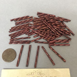 May include: A pile of approximately 50 small, red, glass beads. The beads are about 1 inch long and have a twisted, faceted design. The beads are scattered on a light brown surface.