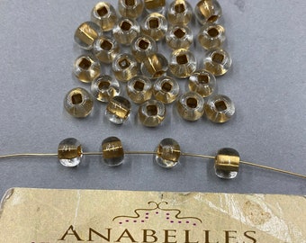 May include: A pile of clear glass beads with gold accents. The beads are round and have a square shape in the center. The beads are arranged in a pile on a white surface.