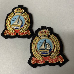 Può includere: Due toppe ricamate nere con filo dorato. Ogni toppa presenta una corona, una barca a vela su sfondo blu e le parole "YACHT CLUB" in filo dorato.