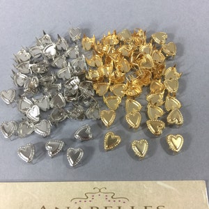 May include: A large pile of silver and gold heart-shaped metal studs. The studs have a raised edge around the heart shape. The studs are arranged in a pile on a gray surface.