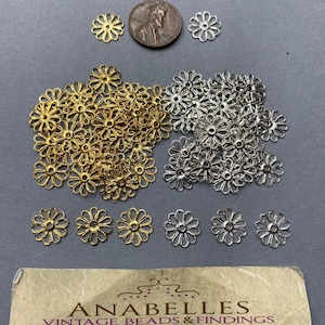 Puede incluir: Un montón de abalorios de flores de metal dorado y plateado. Los abalorios son del tamaño de una moneda de un céntimo de euro. Hay 6 abalorios individuales en la fila inferior, 3 dorados y 3 plateados.