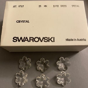May include: A box of clear crystal snowflake charms, labeled "Swarovski" and "Made in Austria". The box is white with black text and the charms are 25 mm in size. The charms are arranged in a pattern on a white surface.