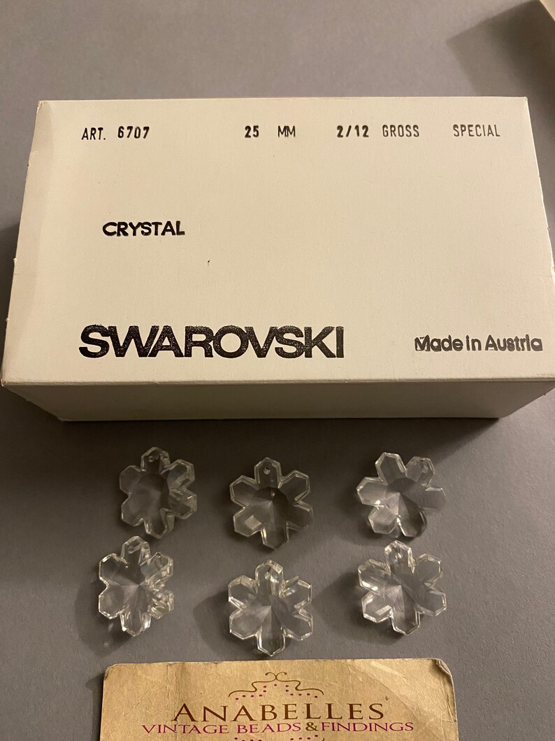 May include: A box of clear crystal snowflake charms, labeled "Swarovski" and "Made in Austria". The box is white with black text and the charms are 25 mm in size. The charms are arranged in a pattern on a white surface.