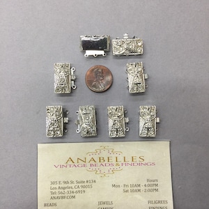 Könnte beinhalten: Sieben silberfarbene Metallverschlüsse mit klaren Strasssteinen. Die Verschlüsse sind rechteckig und haben ein dekoratives Design. Die Verschlüsse sind in einer Reihe auf einer weißen Oberfläche angeordnet. Die Visitenkarte von Anabelles Vintage Beads & Findings befindet sich in der rechten unteren Ecke des Bildes.