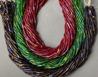 May include: Three strands of iridescent glass beads in shades of red, green, and purple. The beads are rectangular in shape and have a glossy finish. The beads are arranged in a loose circle and are displayed on a white surface. The image includes the text "ANABELLES VINTAGE BEADS & FINDINGS".