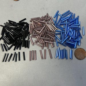May include: An overhead shot displays three piles of small, cylindrical beads in black, rose, and blue. A penny is next to the black beads, and another is near the blue beads. The beads are likely for jewelry making or other crafts.